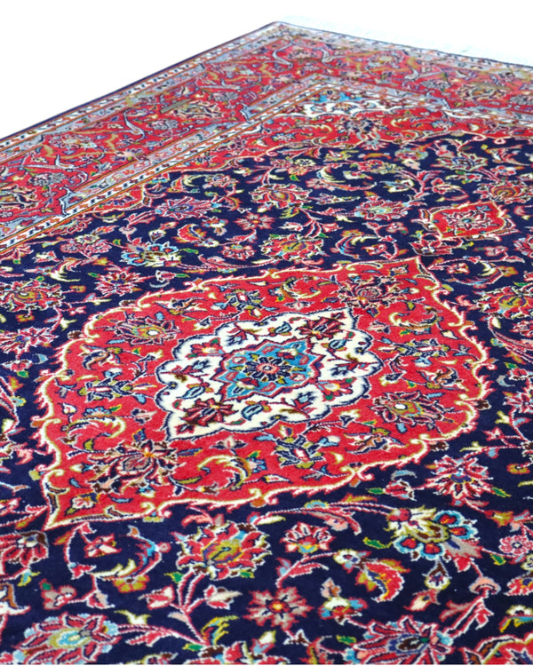 Tappeto Kashan persiano 225x142 cm, annodato a mano, fondo blu notte con motivo floreale e cornice rossa decorata, lana mista seta, certificato di autenticità incluso.
