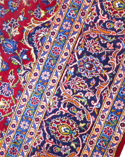 Tappeto persiano Kashan 400x290 cm, fondo rosso con medaglione centrale blu e avorio, cornice scura floreale, annodato a mano in lana vegetale.