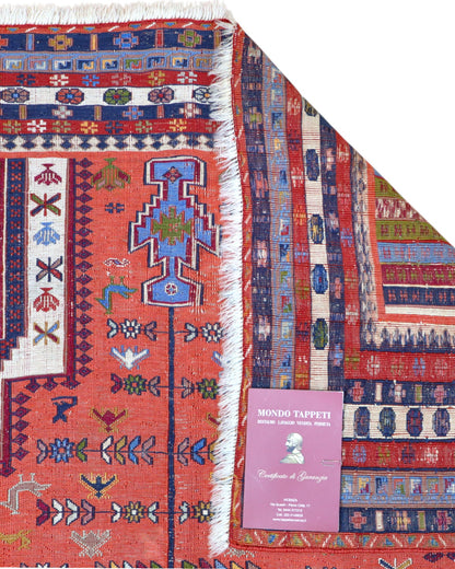 Tappeto Kilim 180x125 cm persiano tessuto a mano, con motivi geometrici tribali e medaglione centrale su fondo rosso mattone.